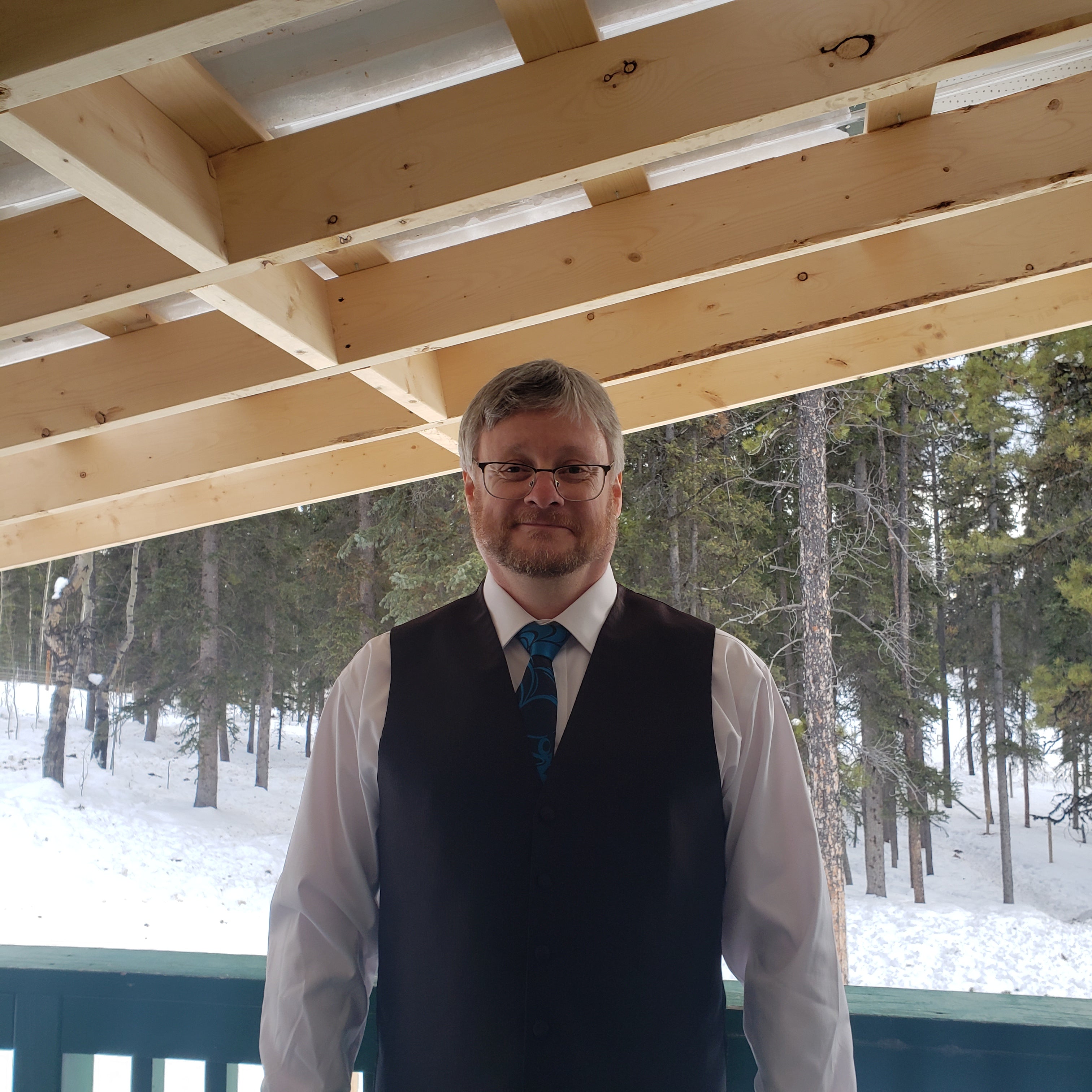 James, co-founder and Owner of Feral as Fhawk™, smiling on a snowy deck surrounded by pine trees, dressed in a white shirt, teal tie, and black vest.